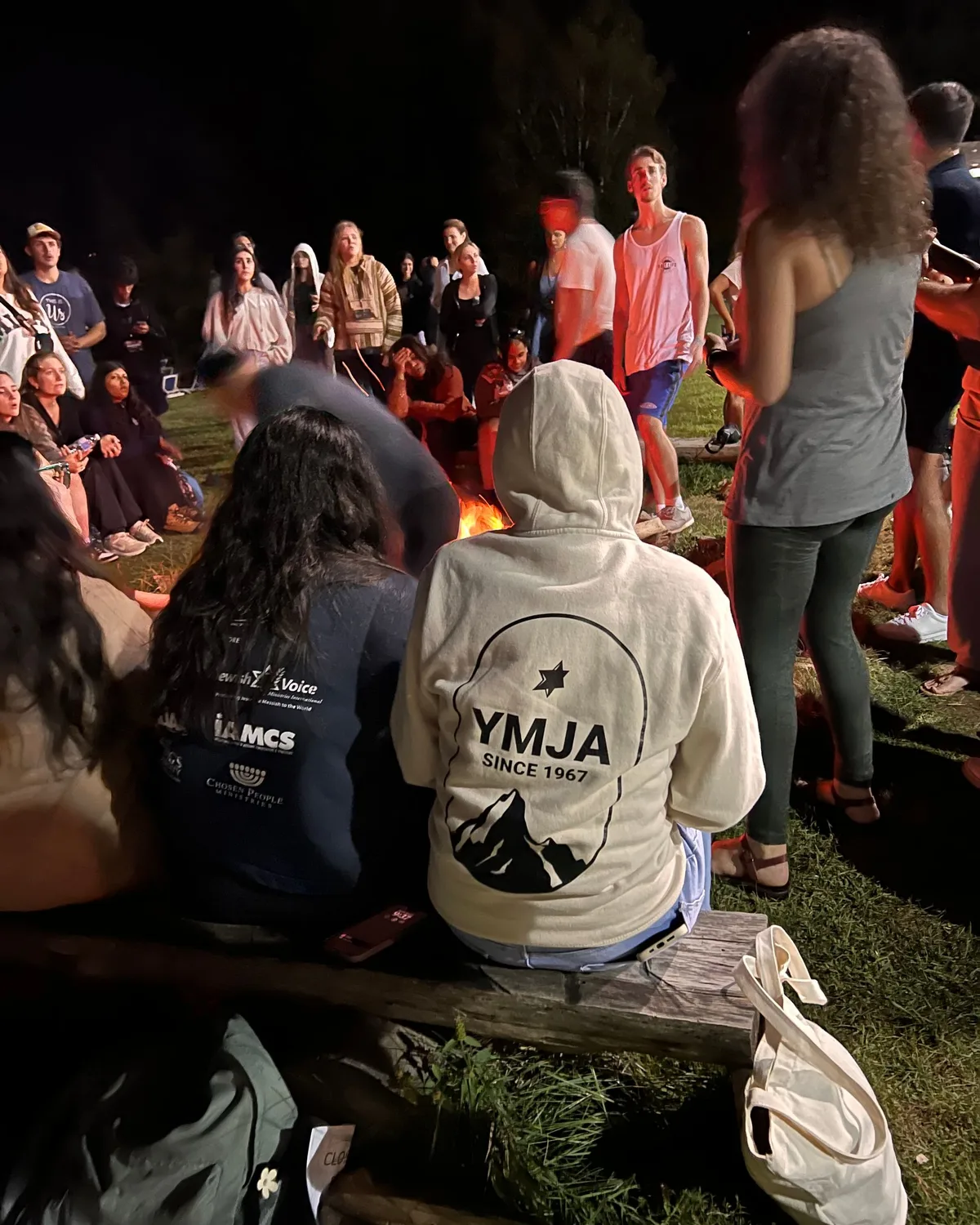 YMJA young people gathered around a campfire at night, one wearing a YMJA Since 1967 hoodie